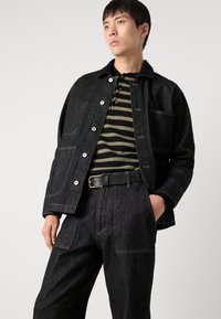 Jachetă din denim negru cu buzunare frontale, asortată cu un tricou polo în dungi negre și oliv. Curea neagră cu cataramă metalică, croială lejeră.