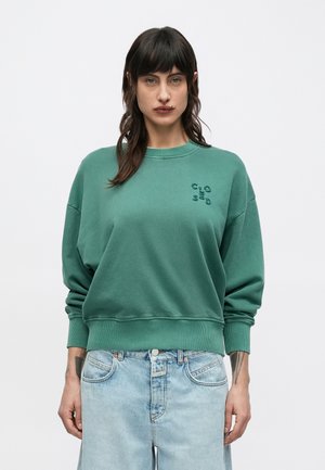 Femeie cu păr închis la culoare, purtând un pulover verde cu un mic detaliu cu pattern pe piept și blugi albaștri deschis, pe un fundal simplu.