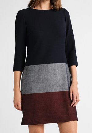 Vrouw draagt een color-block jurk met een zwart bovenstuk, grijze middenstuk en donkerrode rok, met driekwartmouwen en een ronde halslijn.