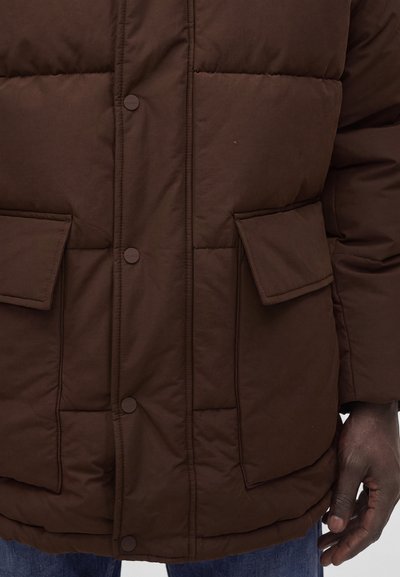 Veste matelassée marron avec boutons-pression et poches à rabat portée par une personne à la peau foncée et un jean bleu.