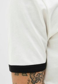 Lähedalt vaade valgele lühikese varrukaga särgile, mille varruka äärel on must äär, ja särgi all on nähtavad tätoveeringud käel.