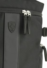 Sac à dos noir en tissu texturé avec un patch en cuir lisse arborant un logo. Fermetures à double zip avec des accessoires en argent.
