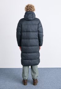 Lång svart puffjacka med huva, quiltat mönster och rak silhuett, tillsammans med lösa ljusgröna byxor och bruna skor.