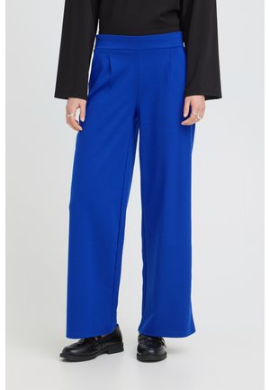 Persona con pantalones anchos de color azul brillante combinados con un top negro de manga larga y zapatos de cuero negros, de pie sobre un fondo liso.