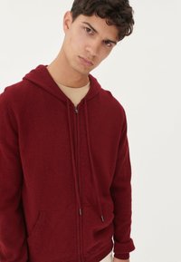 Burgundy zip-up sweatjacka tillverkad av mjukt, strukturerat tyg med dragsko, framfickor och ribbade ärmslut.