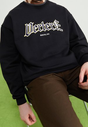 Person sitzt auf einem Stuhl und trägt ein schwarzes Sweatshirt mit den Aufschriften "Vertex" und "Berlin" sowie braune Hosen vor einem grünen Bodenhintergrund.