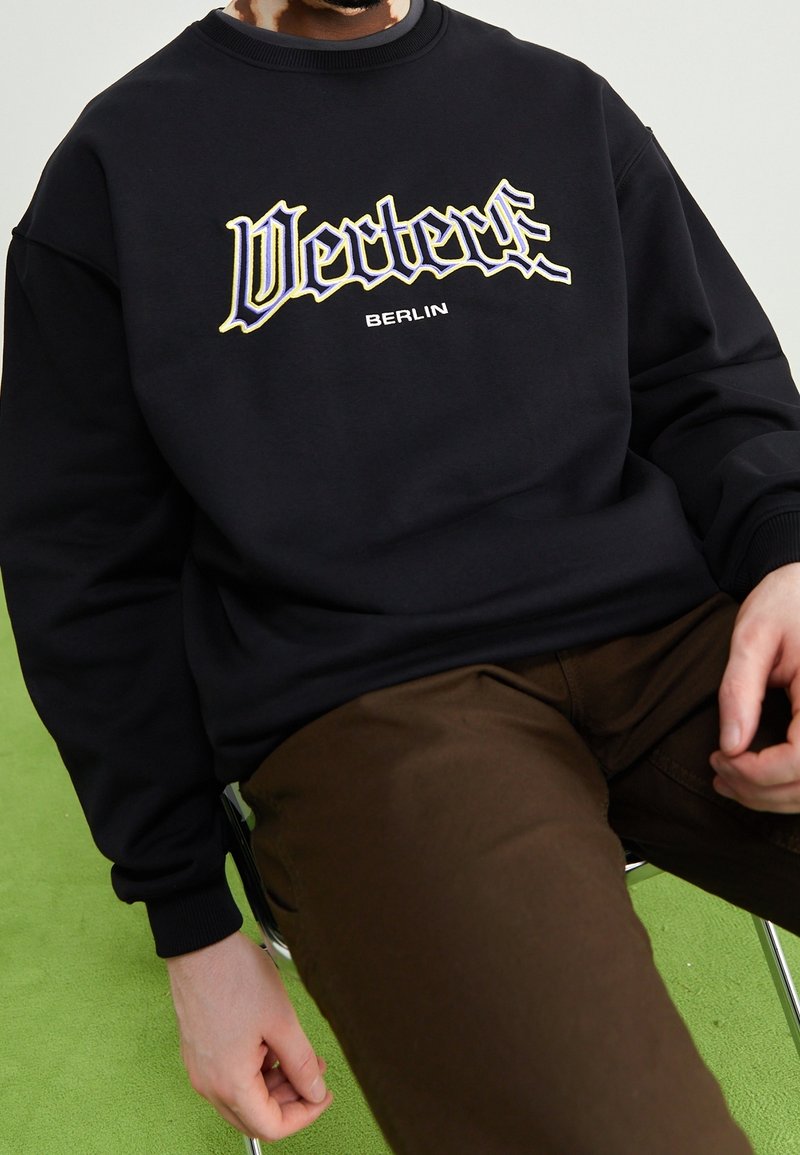 Person seated on a chair wearing a black sweatshirt with "Vertex" and "Berlin" text and brown pants on a green floor background.