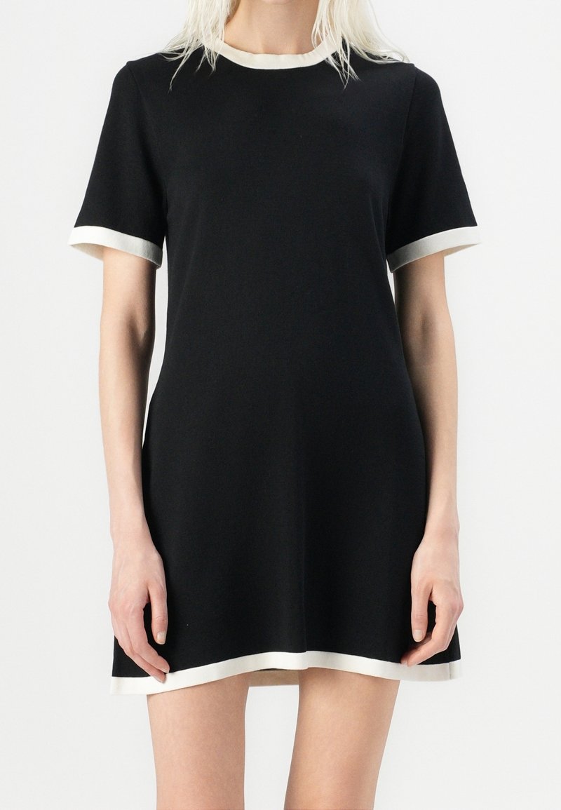 Femme portant une robe courte noire avec un bord blanc au col, aux manches et à l'ourlet sur un fond clair uni.