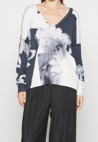 Sweater à manches longues, en V, de couleur marine et blanche avec un imprimé floral ; texture douce, épaules tombantes et bords côtelés. Associé à un pantalon plissé noir.