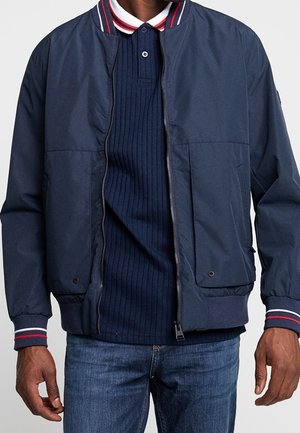 Mann trägt eine marineblaue Jacke mit Reißverschluss über einem marineblauen, gerippten Poloshirt mit rotem, weißem und marineblauem gestreiftem Kragen und Bündchen, kombiniert mit blauen Jeans.