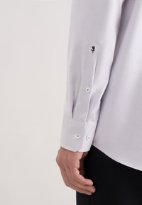 Primo piano di un braccio di una persona che indossa una camicia bianca a maniche lunghe con texture, con polsino abbottonato e un piccolo fiore nero ricamato vicino al gomito.