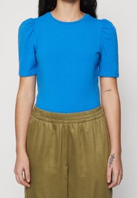 Ljusblå, kortärmad topp med puffade axlar och rund halsringning; kombinerad med ljusgröna byxor med elastisk midja. Slät bomullstextur.