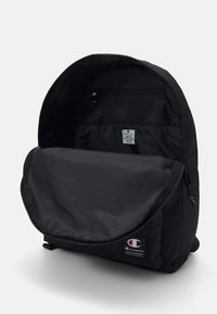 Sac à dos noir avec une ouverture zippée, offrant un intérieur spacieux et une petite poche interne. Fabriqué en tissu durable, avec un détail de logo.