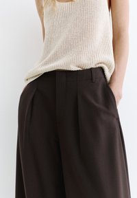 Femme portant un pantalon ample marron foncé taille haute avec plis et un top en tricot beige sans manches, mains dans les poches, sur un fond uni.