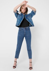 Jeansjakke og høyhåndede jeans i mellomblått. Svart topp under, med sandaler. Jakken har sølvfarget hardware og rullede ermer.