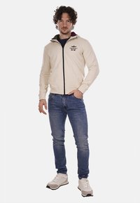 Giacca a zip color crema con dettagli neri e logo, abbinata a jeans blu e sneakers chiare. Il materiale è liscio, con un design aderente.