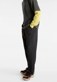 Pantalon noir à coupe décontractée associé à un t-shirt gris foncé et une chemise jaune à manches longues, avec un texte graphique et des baskets multicolores vibrantes.