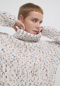 Pull en maille épaisse de couleur crème avec un col roulé haut, présentant des éclats multicolores en bleu, orange et violet.