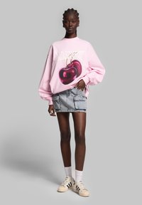 Felpa over-size rosa con grafica di ciliegia, abbinata a una mini gonna in denim chiaro e sneakers beige con strisce nere.