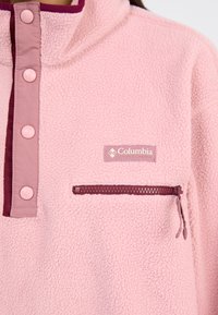 Pull en polaire rose clair avec un col montant, comportant des boutons d'accent violets et une poche zippée, ainsi qu'un patch du logo Columbia.