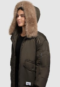 Jachetă pufosă de culoare verde măslinie, cu o glugă mare din blană sintetică, moale și călduroasă, cu buzunare cu fermoar și detalii de design matlasat. Eticheta de marcă este vizibilă.