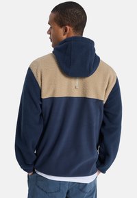 Heren hoodie fleece trui in marineblauw en beige. Heeft een gestructureerde fleece stof, een contrasterend kleurblokontwerp en een ontspannen pasvorm.