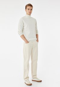 Suéter de cuello alto beige claro combinado con pantalones crema. El calzado presenta un diseño de caña baja con una suela beige. Los detalles incluyen puños acanalados.
