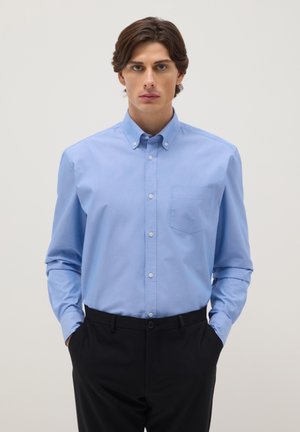 Junger Mann mit mittellangen braunen Haaren trägt ein hellblaues Hemd, das in schwarze Hosen gesteckt ist, Hände in den Taschen, neutraler Hintergrund.