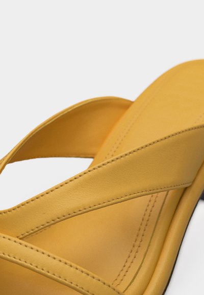 Κίτρινο δερμάτινο σανδάλι με φαρδιές διασταυρωμένες τιράντες και εμφανές ράψιμο σε λευκό φόντο.