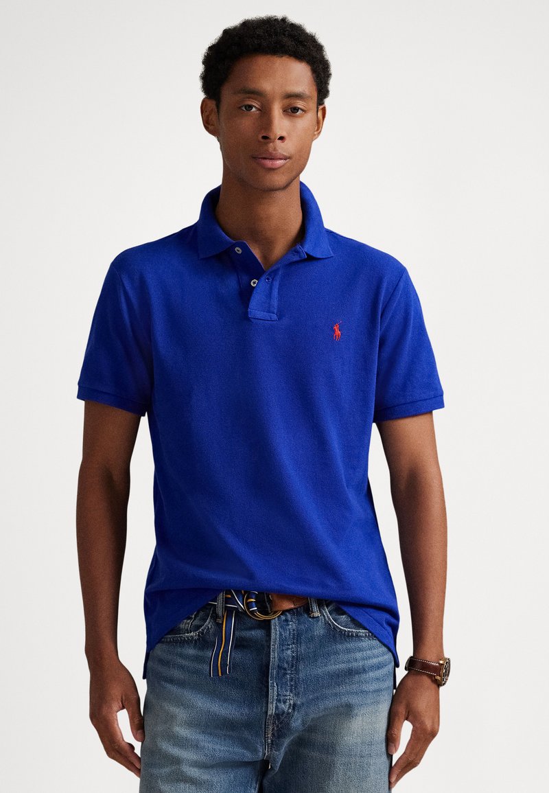 Polo azul royal com pala de dois botões e logótipo vermelho bordado no peito; usado com jeans azuis desbotados e cinto padrão.