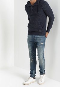 Felpa con cappuccio testurizzata blu navy, con tasca frontale, lacci e due bottoni; abbinata a jeans chiari strappati e sneakers bianche.