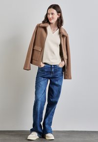 Braune Wollmischgewebe-Jacke mit einem plüschigen Kragen, beigem V-Ausschnitt-Oberteil und blauen weiten Jeans, kombiniert mit cremigen Sneakers vor einem neutralen Hintergrund.