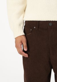 Pantalones de pana marrones con estrías verticales, combinados con un suéter de punto crema. Los detalles visibles de los bolsillos y los botones realzan el estilo.