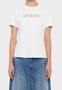 Vit bomulls-t-shirt med guldfärgade glitterbokstäver som lyder "ARMANI EXCHANGE", med klassisk rund halsringning och korta ärmar.