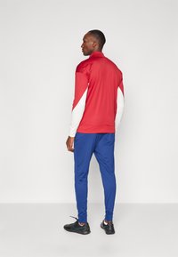 Red track jacket with white accents, paired with blue pants and black athletic shoes. Smooth fabric, slim fit, and long sleeves.