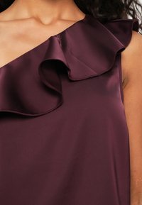 Burgundy topp med en axel-design och en volangdetalj. Slät satinkvalitet, åtsittande silhuett och inga synliga mönster eller detaljer.