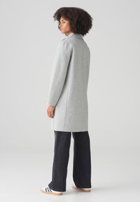 Grijze wollen mix jas met een kraag, zijsplitsen en lange mouwen, gecombineerd met donkere flare jeans en witte sneakers met blauwe strepen.