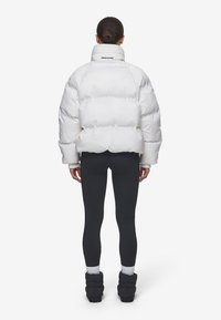 Vitt pufferjacka med hög krage, med en glänsande yta och kviltat mönster. Svarta leggings och ribbmuddar fulländar outfiten.