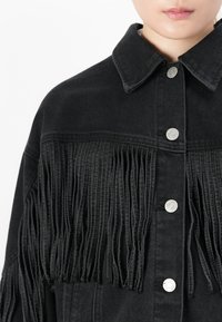 Primo piano di una giacca di denim nera con dettagli in frange verticali e bottoni a pressione argentati sul davanti, indossata da una persona.