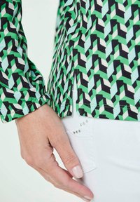 Grün-schwarz gemusterte Bluse mit geometrischem Design, mit hohem Kragen und gerafften Manschetten; weiße Hose mit Strassakzenten.