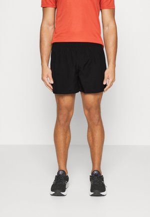 Mann trägt schwarze Sportshorts, schwarze Laufschuhe und ein orangefarbenes kurzärmeliges Shirt, steht vor einem einfarbigen Hintergrund.