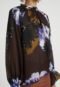Durchsichtige, langärmelige Bluse mit einem Rüschenkragen. Verfügt über abstrakte Blumenmuster in Blau, Weiß und Braun auf einem dunklen Hintergrund.