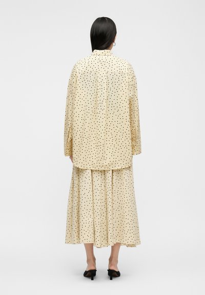 Femme aux cheveux noirs raides portant une chemise à manches longues crème et une jupe plissée assortie à pois noirs, debout sur un fond gris.