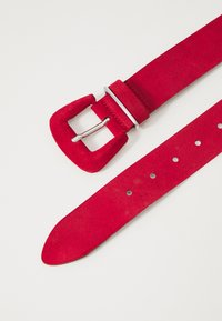 Cintura in suede rosso con fibbia rettangolare. Presenta una texture liscia e diversi fori di regolazione lungo la cintura. Accentuata con hardware argentato.