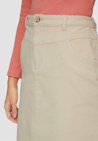 Jupe en velours côtelé beige avec poche avant et taille boutonnée portée avec un haut corail à manches longues, montrant la main gauche détendue sur le côté.