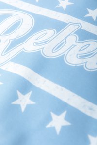Lys blått stoff med hvite stjerner og teksten "Rebel" i en tykk, litt slitt font på midten. Jevn tekstur.