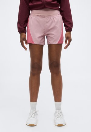Person iført lyserøde træningsshorts med mørkere lyserøde sidepaneler, hvide sokker, hvide sneakers og en vinrød langærmet top, stående oprejst.