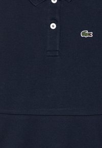 Tengerészkék póló, texturált anyagból, három fehér gombbal, és egy kis zöld krokodil logóval hímzett a mellrészen.