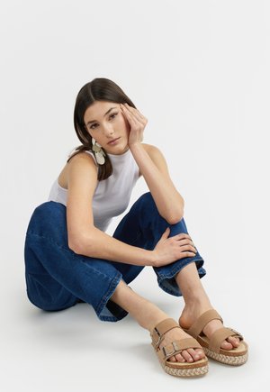 Giovane donna seduta a terra con top bianco senza maniche, jeans blu, sandali beige e grandi orecchini bianchi, appoggiando la testa sulla mano.