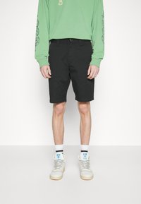 Shorts noirs arrivant au genou avec une coupe décontractée, en tissu mélangé coton. Associés à un maillot à manches longues vert et des baskets blanches montantes.